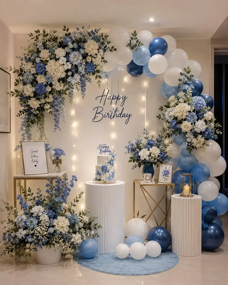 Elegant blue and white birthday party decoration featuring floral arrangements, balloon garlands, a birthday cake, candles, and illuminated backdrop.