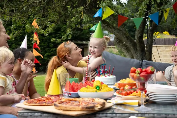 Children celebrating a birthday outdoors with colorful party hats, balloons, bunting, birthday cake, pizza, fruit, and snacks at a picnic table.